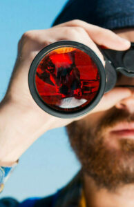 Opening of new horizons. Handsome young bearded man carrying backpack and looking through binoculars at camera